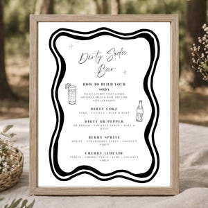 May include: A framed sign with the text "Dirty Soda Bar" in elegant script. The black and white sign includes instructions for making soda drinks, with illustrations of a glass and a bottle. The frame is a light brown color.
