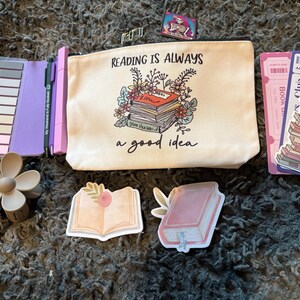 May include: An assortment of book-themed accessories, including a canvas pouch with the text "Reading is Always a Good Idea," stickers, bookmarks, and a flower-shaped object. The items are arranged on a textured surface.