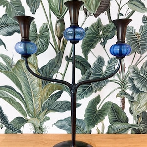May include: A black metal candelabra with three arms, each holding a dark brown candle cup and a blue glass orb. The candelabra stands on a round base. The background features a green and white botanical wallpaper.