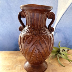 Könnte beinhalten: Eine braune, geschnitzte Holzvase mit zwei verzierten Griffen. Die Vase hat detaillierte Schnitzereien und einen breiten Sockel. Der Hintergrund ist hellblau und die Vase steht auf einer Holzoberfläche. Rechts von der Vase befindet sich eine kleine Pflanze.