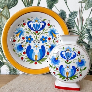 Peut inclure: Un ensemble d'assiette et de vase en céramique blanche avec un motif floral bleu, jaune et rouge. L'assiette a un bord jaune et le vase a une base rouge. Le motif floral comprend des fleurs bleues stylisées et des feuilles vertes.