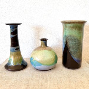 May include: Three ceramic vases with a glossy finish. The vases feature a color palette of brown, blue, green, and yellow. The vase on the left is tall and flared, the middle vase is round, and the vase on the right is cylindrical.