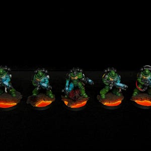 May include: Five miniature figures of soldiers in green armour with blue accents, each holding a futuristic weapon. They stand on bases with a lava-like orange glow, set against a black background.
