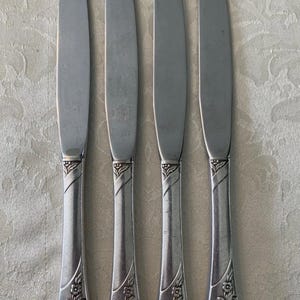 May include: Four stainless steel knives with a floral design on the handles. The blades are smooth and curved, and the handles have a decorative pattern near the base. The knives are arranged on a light-colored surface.