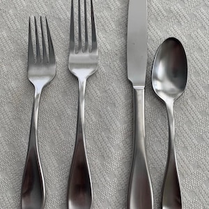 Puede incluir: Un juego de cuatro cubiertos de acero inoxidable: dos tenedores, un cuchillo y una cuchara. La cubertería tiene un diseño sencillo y elegante con mangos redondeados y un acabado mate. Los utensilios están dispuestos sobre una superficie texturizada de color gris claro.