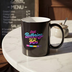 Puede incluir: Taza de café negra con el texto "Still Buffering from the 90s" en una fuente colorida y retro. La taza tiene un interior blanco y un asa negra. En el fondo se ven una pizarra de menú y un azucarero.