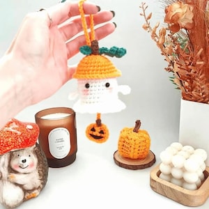 May include: A crocheted ghost ornament wearing an orange hat with a green leaf, featuring a small dangling jack-o'-lantern. The ghost has black eyes and pink cheeks. A separate small crocheted orange pumpkin sits on a wooden slice nearby.