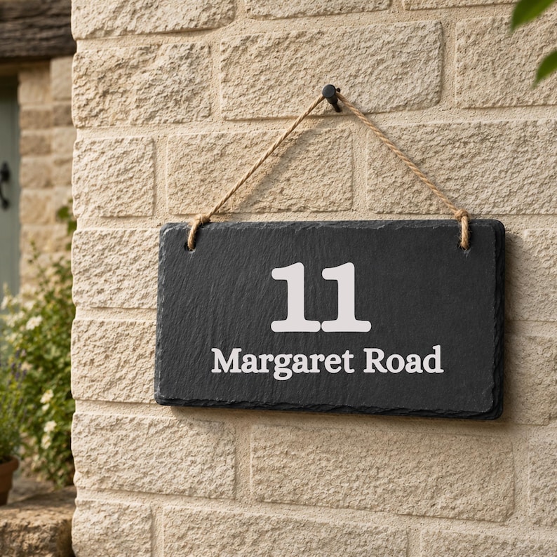 Puede incluir: Un letrero rectangular de pizarra negra para casa que muestra el n&uacute;mero 11 y el texto Margaret Road en letras blancas. El letrero est&aacute; suspendido por una cuerda gruesa de yute color canela sujeta a un gancho de metal negro en una pared de ladrillo.