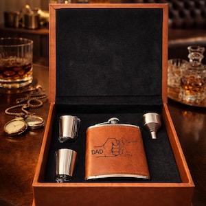 May include: A brown leather flask set presented in a box. The set includes a flask with the word "DAD" and two fists engraved, two shot glasses, and a funnel. The box is lined with black fabric.