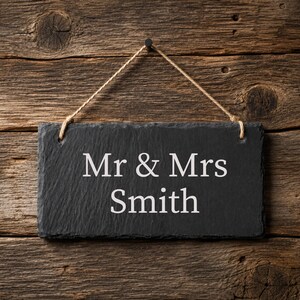 Puede incluir: Un letrero rectangular de pizarra negra con bordes rugosos y texturizados, colgado de una cuerda fina de yute. El letrero presenta el texto grabado en blanco "Mr & Mrs Smith" centrado en su superficie, mostrando el nombre de forma clara.