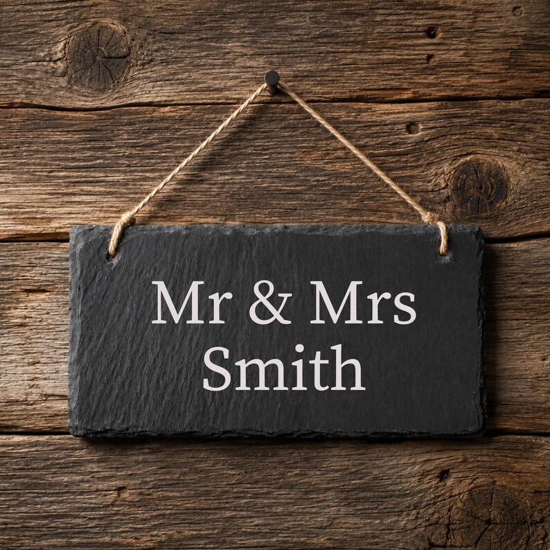 Puede incluir: Un letrero rectangular de pizarra negra con bordes rugosos y texturizados, colgado de una cuerda fina de yute. El letrero presenta el texto grabado en blanco "Mr & Mrs Smith" centrado en su superficie, mostrando el nombre de forma clara.