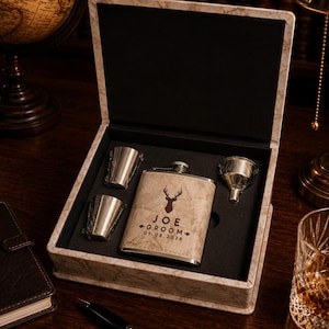 May include: A beige marble-patterned gift box containing a matching metal flask, two small metal shot glasses, and a metal funnel. The flask is engraved with a deer head silhouette, the text JOE GROOM, and the date 01.08.2026 in dark lettering.