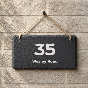 Puede incluir: Un letrero rectangular de pizarra gris oscuro para casa con el n&uacute;mero 35 y el texto Wesley Road impresos en blanco. El letrero tiene una textura de piedra natural rugosa y est&aacute; suspendido por una cuerda de yute color canela a un gancho negro.