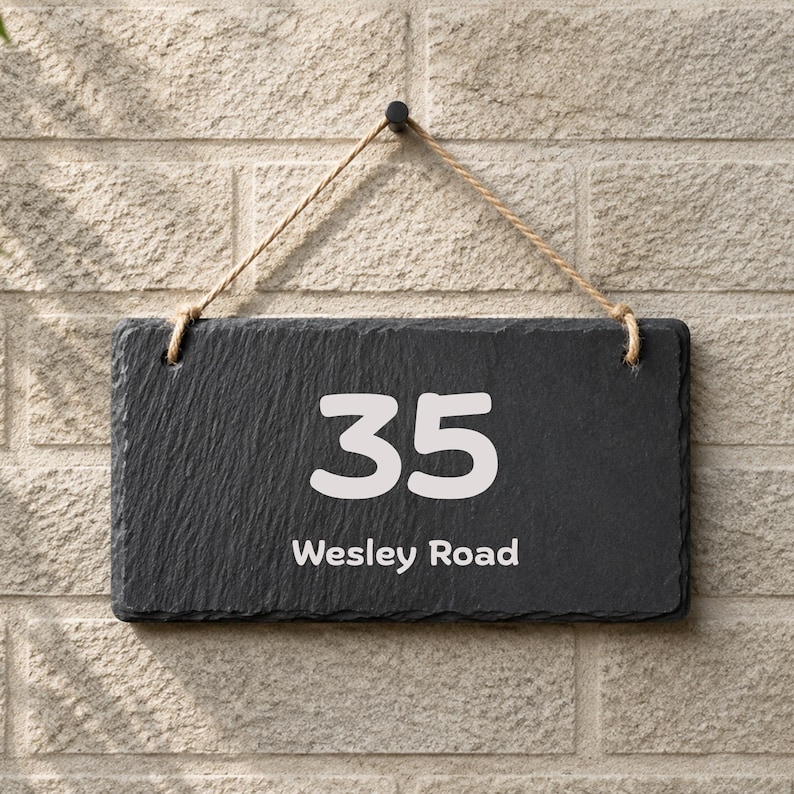 Puede incluir: Un letrero rectangular de pizarra gris oscuro para casa con el n&uacute;mero 35 y el texto Wesley Road impresos en blanco. El letrero tiene una textura de piedra natural rugosa y est&aacute; suspendido por una cuerda de yute color canela a un gancho negro.