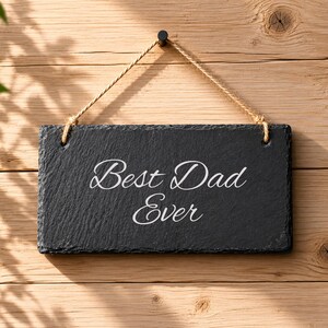 Puede incluir: Un letrero rectangular de pizarra negra con bordes rugosos y texturizados. El texto "Best Dad Ever" est&aacute; grabado en el centro con una fuente cursiva blanca y elegante. El letrero cuelga de una cuerda de yute natural a trav&eacute;s de dos agujeros.