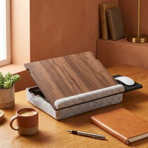 May include: A wooden laptop desk with a gray padded base and a small black mouse pad. A white mouse sits on the pad. A brown mug, a pen, and a leather-bound notebook are also on the desk. The desk is on a wooden surface.