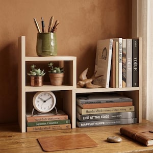 May include: A light-colored wooden desk organizer with multiple shelves. It holds books, a clock, potted plants, pens, and decorative items. The books include titles like "Vogue" and "Handcrafted: A History of Object Making."