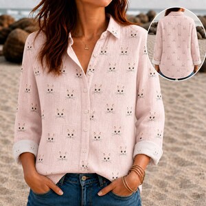 May include: A light pink long-sleeve button-down shirt featuring a repeating pattern of small cat faces. The shirt has a classic collar, a front button placket, and sleeves with folded cuffs. The back view is shown in a circular inset.