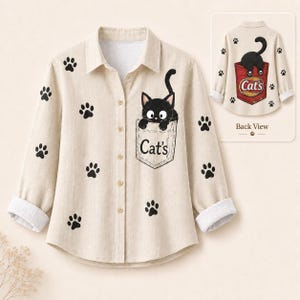 May include: A beige long-sleeve button-down shirt featuring a black cat graphic peeking out of the chest pocket with the text Cat's. The shirt is decorated with black paw prints and includes an inset showing the back view with a cat in a pocket.