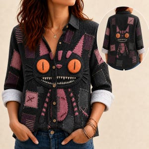 May include: A dark gray long-sleeve button-down shirt featuring a large, stitched cat face graphic with bright orange eyes and a wide, toothy grin. The shirt is decorated with various purple and gray fabric patches and visible stitching details.