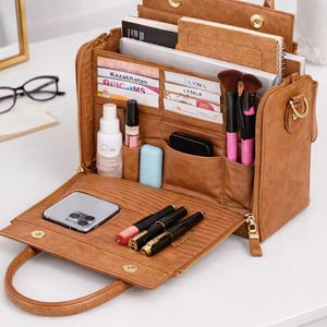 May include: A brown leather organizer bag with multiple compartments, open to reveal contents. Includes notebooks, makeup brushes, pens, a phone, and beauty products. The bag has a top handle and a zipper closure.