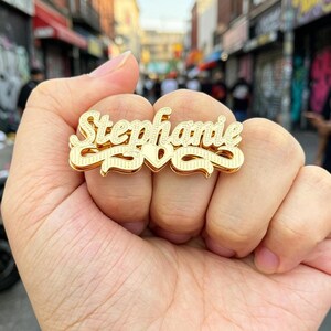 Puede incluir: Un anillo con nombre en tono dorado que presenta el nombre Stephanie en letra cursiva. El diseño incluye una pequeña forma de corazón centrada debajo del nombre y adornos decorativos en ambos extremos, con un acabado metálico texturizado.