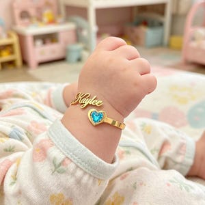 Könnte beinhalten: Ein goldfarbenes, offenes Armband mit dem Namen Kaylee in Schreibschrift an einem Ende und einem kleinen herzförmigen Anhänger mit einem leuchtend blauen Stein am anderen Ende, getragen an einem kleinen Handgelenk über einem weißen Blumenkleidungsstück.
