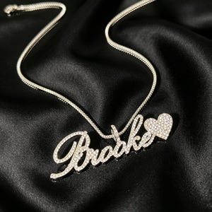 Könnte beinhalten: Eine silberfarbene Halskette mit dem Namen "Brooke" in Schreibschrift, verziert mit glitzernden Kristallen. Ein herzförmiger Anhänger, ebenfalls mit Kristallen besetzt, ist am Ende des Namens befestigt. Die Kette liegt auf schwarzem Satin.