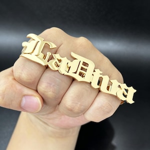 Può includere: Un anello dorato per più dita che riporta il nome LaDiva in un carattere gotico stilizzato. Il pezzo di metallo si estende su quattro dita, con ogni lettera collegata per formare un gioiello continuo con una finitura lucida e riflettente.