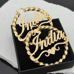 Op de afbeelding: Goudkleurige oorringen met de naam "India" in een sierlijk lettertype. De ringen hebben een gedraaid touwontwerp. Een klein hartje is afgebeeld op een oorbel. De oorbellen worden in een zwarte doos gepresenteerd.