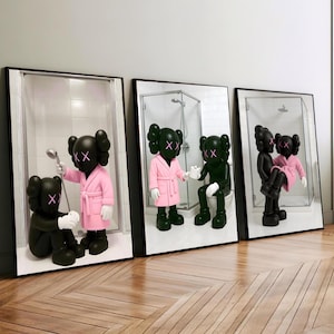 May include: Three framed art prints depicting KAWS figures in bathroom settings. The figures are black and green, wearing pink robes and 'XX' eyes. One figure holds a shower head, another sits on a toilet, and the third is carrying another.