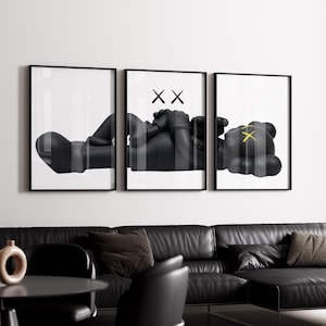 May include: Triptych wall art featuring a black and white abstract figure in a reclining pose. The artwork is framed in black and hangs above a black leather sofa. The figure has an 'XX' symbol on the eyes and hands.