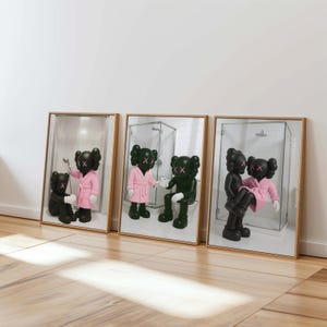May include: Three framed art prints featuring cartoon figures in bathroom scenes. The figures are black with X-shaped eyes and mouths. One figure wears a pink robe. The frames have a light wood finish and are displayed against a white wall.