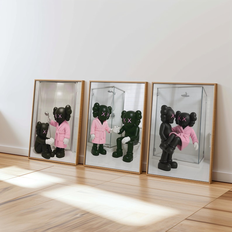 May include: Three framed art prints featuring cartoon figures in bathroom scenes. The figures are black with X-shaped eyes and mouths. One figure wears a pink robe. The frames have a light wood finish and are displayed against a white wall.