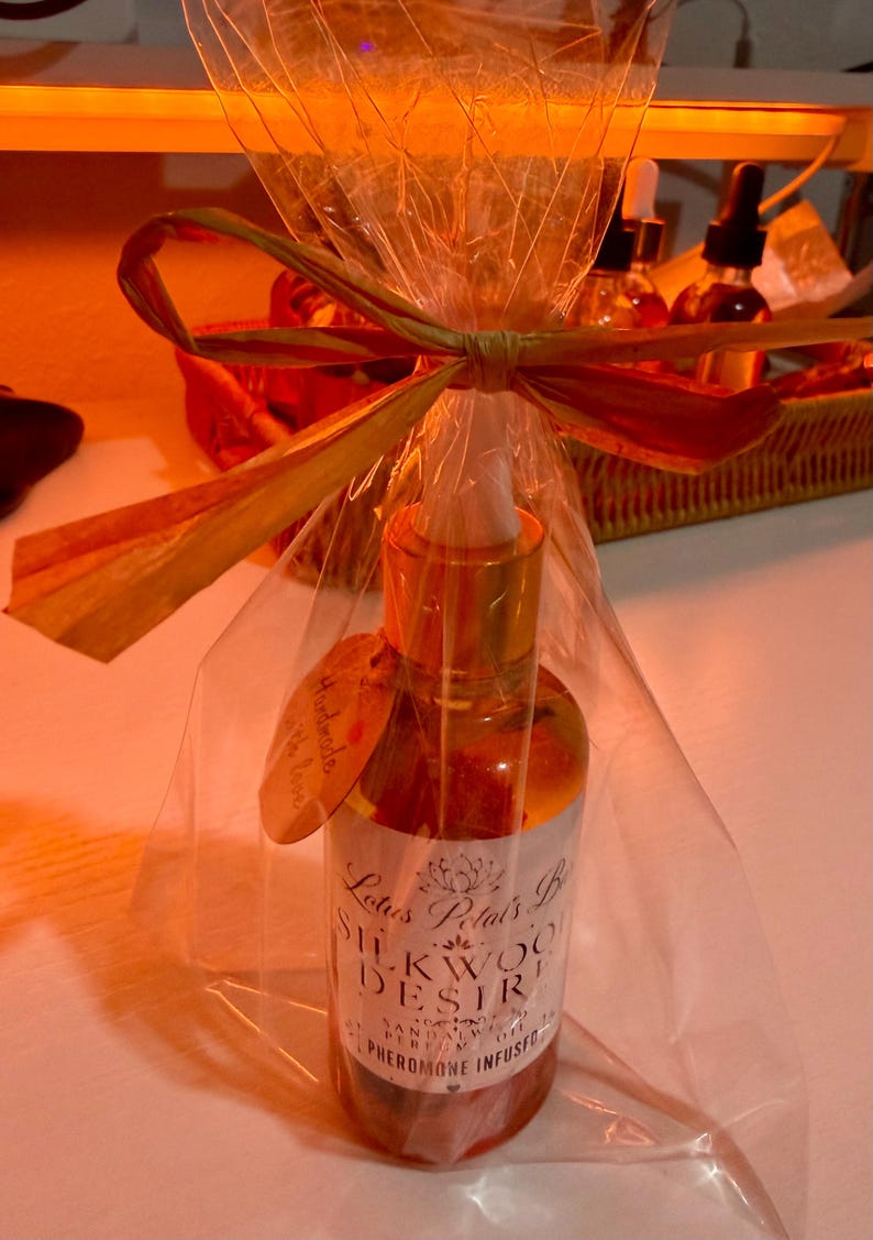May include: A glass bottle with a gold cap and dropper, wrapped in cellophane and tied with a raffia bow. The label reads "Silkwood Desire" and "Pheromone Infused". A small tag says "Handmade with love".