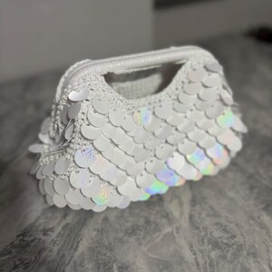 Pode incluir: Uma bolsa de crochê branca com uma estrutura rígida e uma superfície coberta por lantejoulas circulares iridescentes sobrepostas que refletem a luz com um brilho sutil de arco-íris. A bolsa possui uma alça superior e uma textura de malha distinta.