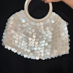 Pode incluir: Uma bolsa branca com lantejoulas e uma alça circular. A bolsa é coberta por lantejoulas redondas e iridescentes, e a alça é de cor bege clara. A bolsa tem formato de semicírculo.