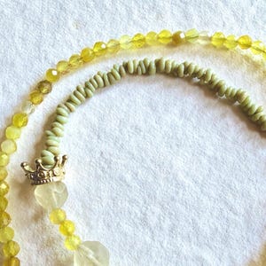 Può includere: Una collana composta da una fila di perline gialle sfaccettate, una sezione di piccole perline cilindriche verde pallido e perline gialle traslucide più grandi. Il gioiello include un ciondolo a corona dorato e una chiusura a gancio e anello.