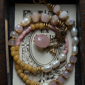Puede incluir: Un collar que presenta una mezcla de perlas de agua dulce blancas, piedras de color rosa pálido facetadas y cuentas amarillas mate, acentuado con espaciadores de metal dorado y un colgante central de piedra rosa facetada con un dije de mano.