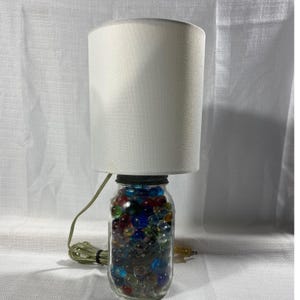Puede incluir: Una lámpara de mesa decorativa con una pantalla cilíndrica blanca. La base es un tarro de cristal transparente lleno de canicas de colores. La lámpara tiene un cable verde y un enchufe estándar.