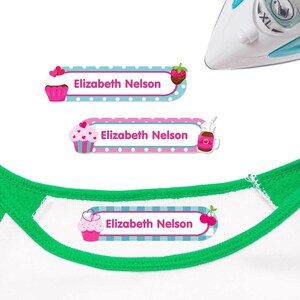 Puede incluir: Tres etiquetas de nombre personalizadas con diseños de cupcake y fresa. Las etiquetas tienen una combinación de colores rosa y azul con el nombre "Elizabeth Nelson" impreso. Una etiqueta está en una camiseta blanca con cuello verde.