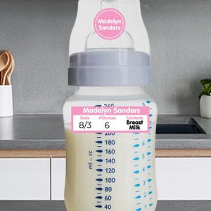 May include: A clear baby bottle with a light blue cap and a pink label that reads "Madelyn Sanders." The label indicates the contents are breast milk, with a date of 8/3 and 6 ounces. The bottle has measurements in milliliters and ounces.