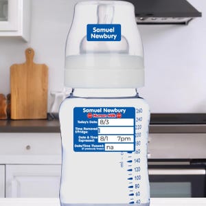 May include: A clear baby bottle with a white top and blue labels. The labels read "Samuel Newbury" and include dates and times for expressed human milk. The bottle has a scale in milliliters and is in a kitchen setting.