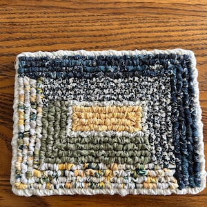 May include: A handmade, rectangular coaster with a patchwork design. The coaster features a variety of fabrics in shades of blue, green, yellow, and white, arranged in a geometric pattern. The edges are finished with a white border.