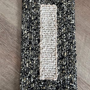 Puede incluir: Una alfombra negra y blanca con un diseño rectangular de color crema en el centro. La alfombra está hecha de tejido tejido y tiene una superficie texturizada.