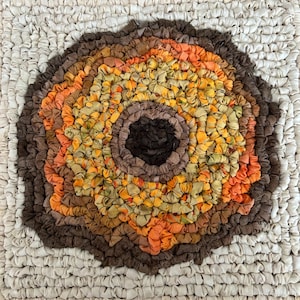 May include: A handmade, square rug with a tree ring design. The center is dark brown, surrounded by rings of orange, yellow, and brown. The outer border is a cream color, creating a rustic, textured appearance. The rug is made of fabric strips.
