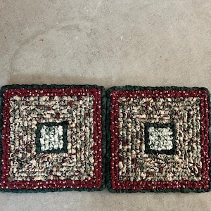 May include: Two square, handmade rag rugs. Each rug features a central square design in shades of green and white, surrounded by a patchwork of red, green, and beige fabrics. The edges are finished with a dark green border.