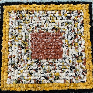 May include: Handmade square trivet with a patchwork design. The center features a textured, rust-colored square. The trivet is surrounded by layers of fabric in yellow, black, and light blue, creating a decorative and functional item for the kitchen.