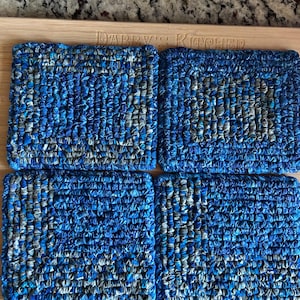 May include: A set of four square coasters crafted from braided fabric strips in shades of blue, navy, and grey. Each coaster features a dense, textured weave pattern with a consistent square shape and uniform size, arranged in a two-by-two grid.