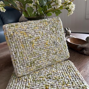 May include: Two rectangular, hand-woven placemats in shades of green, yellow, and white. The placemats have a textured, braided appearance. A vase of white flowers and greenery sits in the background.